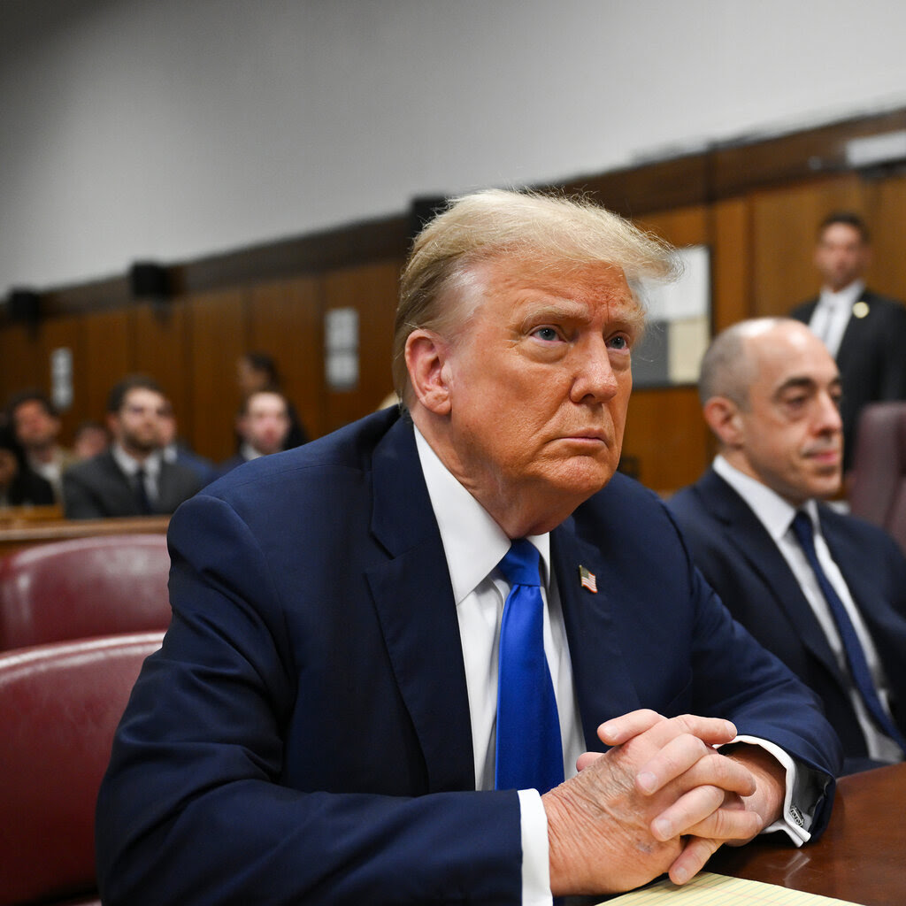 Donald Trump sits at a courtroom table, wearing a blue suit and a bright blue tie, his hands folded in front of him with a yellow legal pad under them