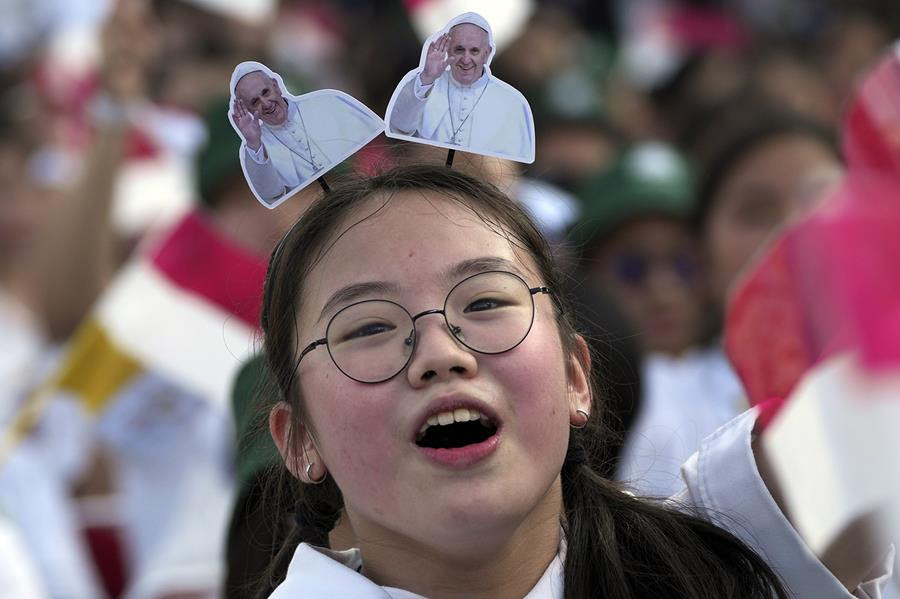 A young Asian girl wearing a headband that has two pictures of Pope Francis waving attached to it looks towards the viewer. There is a blurry crowd of people behind her.