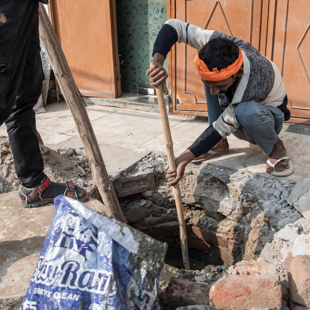 Two men reach poles down into a drain. One of the men is wearing an orange headband.