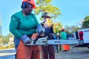 Alexis Powell works with a chainsaw on a sunny day with a team member assisting in the background