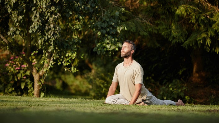 A man is doing a pigeon pose stretch outside in nature.