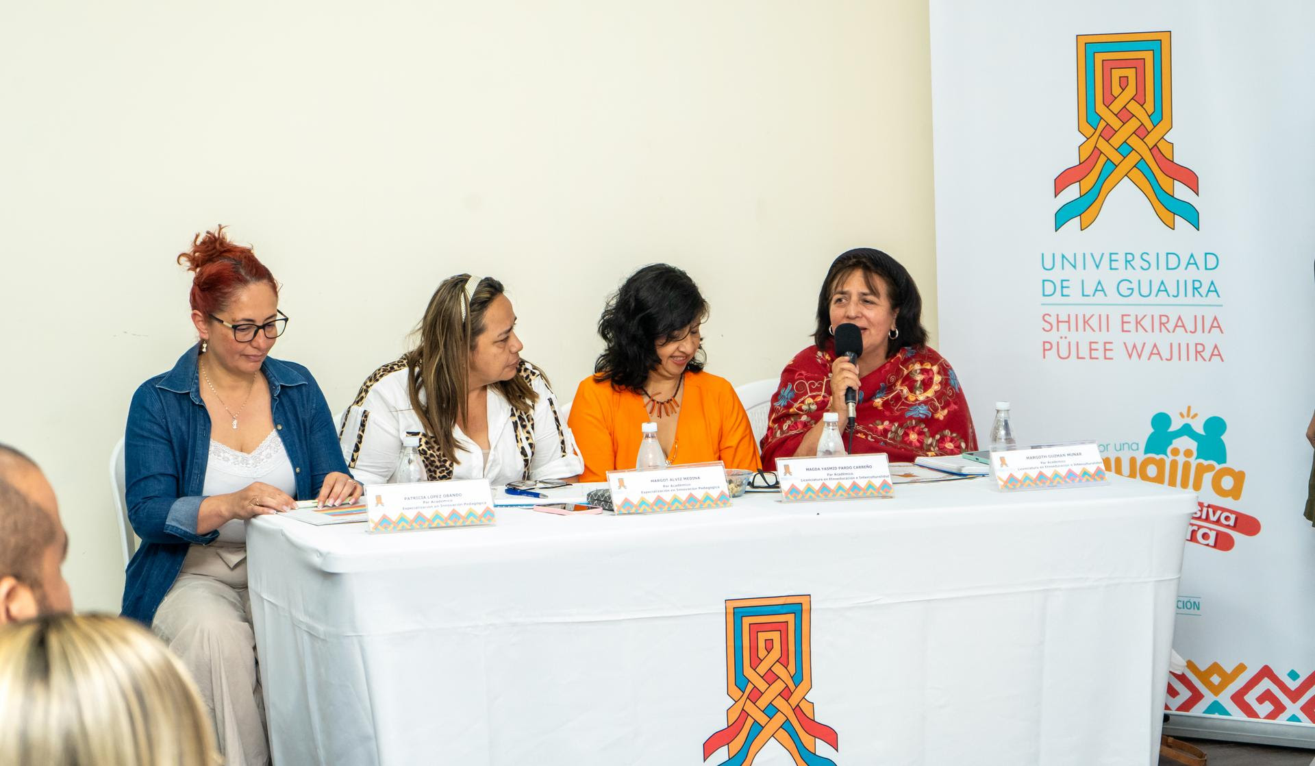 Cuatro mujeres están sentadas en una mesa en una conferencia o panel. Están hablando y mirando a la persona que sostiene un micrófono, quien parece estar interviniendo en la discusión. Detrás de ellas hay un cartel que dice ''Universidad de La Guajira'', con un diseño colorido en la parte superior. Las mujeres están vestidas en tonos vibrantes y formales.