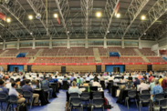 Many people sit at tables set up for a committee meeting in a large sports anex-style building.