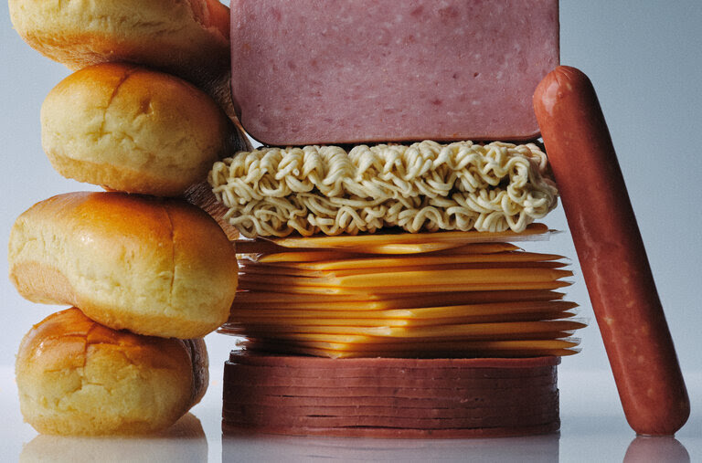 Deli meat, dried ramen noodles, sliced cheese, two hot dogs and hot dog buns in front of a gray background.