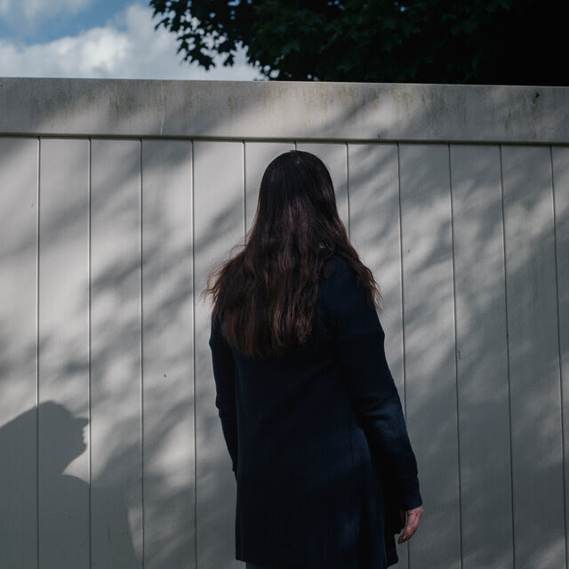 With her back to the camera, Patrice Motz faces a tall, solid fence. She and foliage cast shadows on the gray surface.