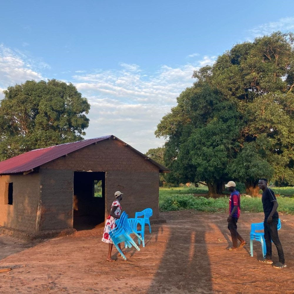 People standing together in front of a small hut, holding plastic chairs.