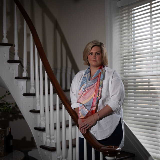 A portrait of Leelee Ray, who wears a white blouse with sleeves rolled halfway up her forearm and a colorful scarf around her neck. She stands on a large curving stair in her home.