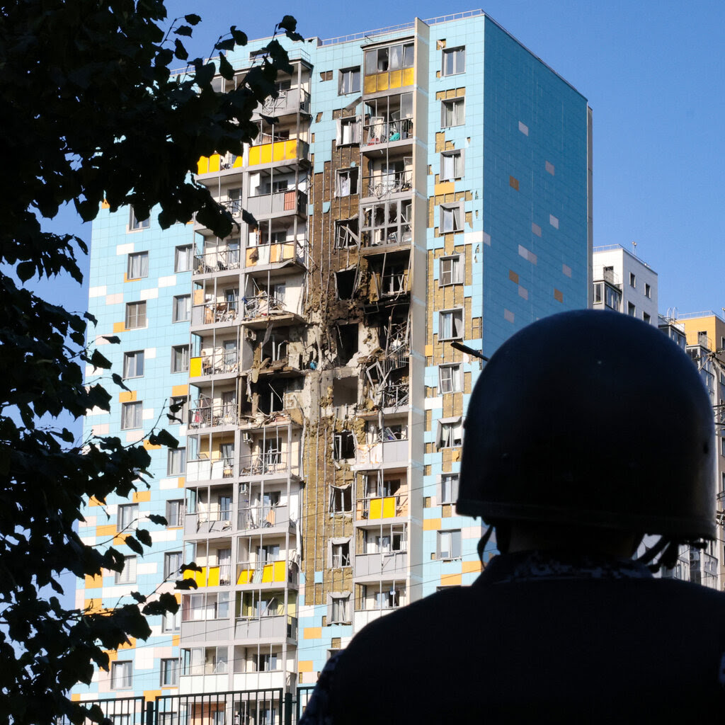 A figure in silhouette wearing a military-style helmet looks up at a damaged high-rise residential building. 