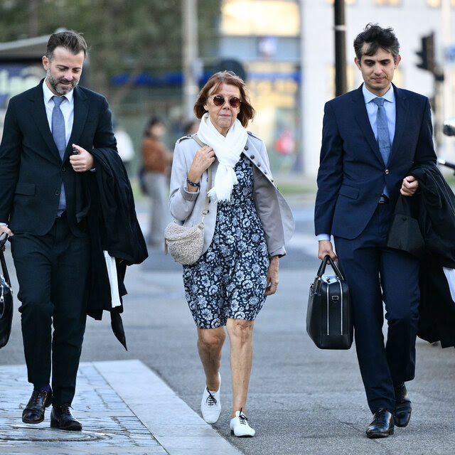 Gisèle Pelicot, wearing a blue, patterned dress along with sunglasses and a white scarf, walks between two men in suits.