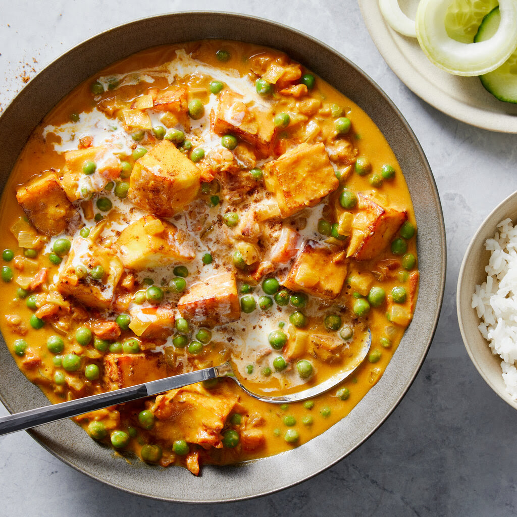 A gray bowl filled with an orange-hued sauce dotted with peas and tofu sits next to a small bowl of rice and a plate of onions and cucumbers.