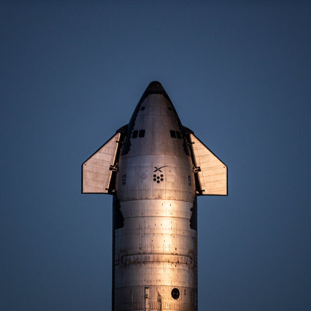 The winged tip of a large rocket appears against a cloudless blue sky at sunset.