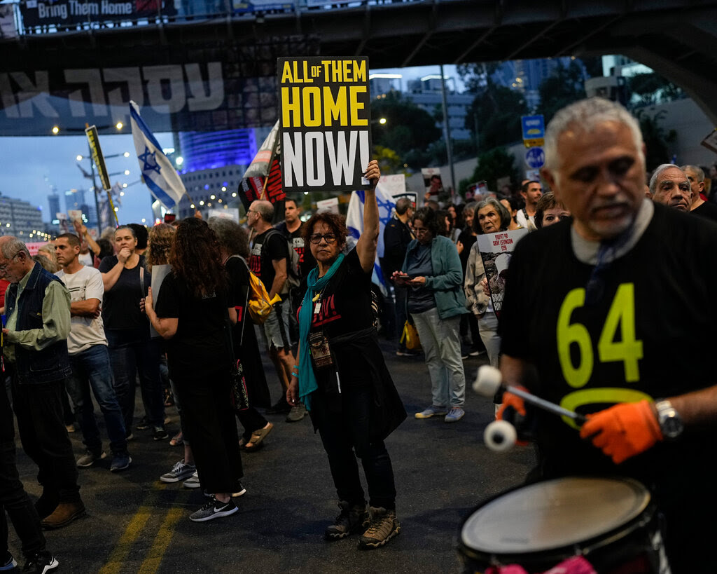 A crowd of demonstrators. Some are waving Israeli flags and a man plays drums. At the center of the photo, a woman holds a sign reading “All of Them Home Now!”