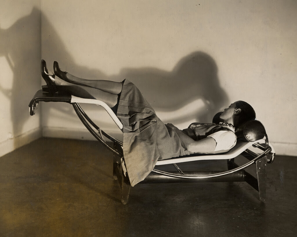 A woman wearing a pleated skirt and a white top reclines in a chaise longue with raised legs and a tubular pillow. A shadow of the chaise longue is projected on the wall.