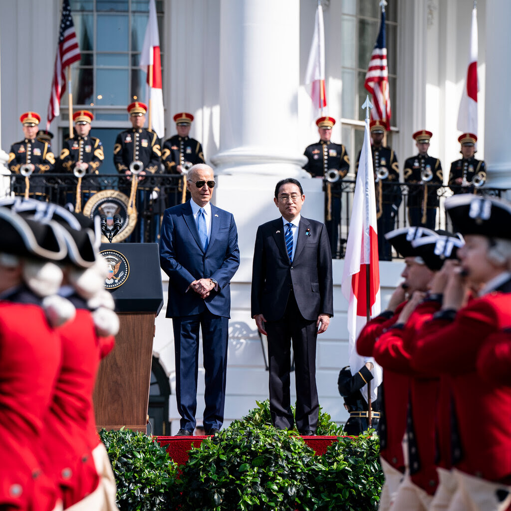 President Biden and Fumio Kishida stand side by side in front of the White House. People in red coats march in front, as people in uniform stand behind them, with U.S. and Japanese flags.