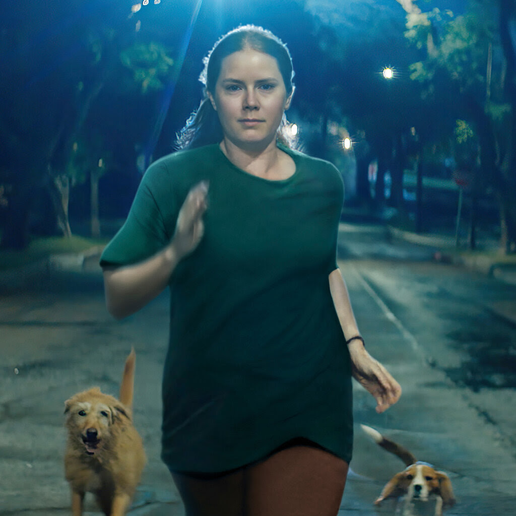 Amy Adams, in a green shirt, runs down a street followed by two dogs.