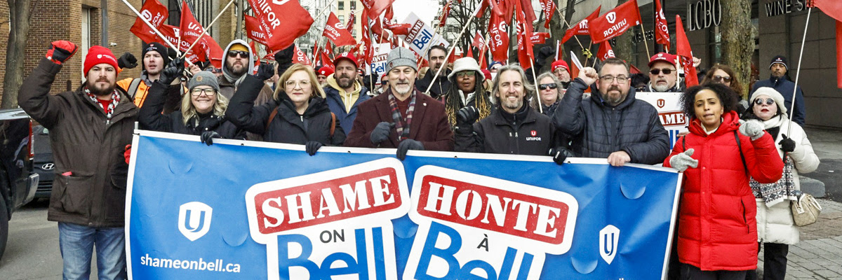 Unifor members marching down a main street waving flags and a large "Shame on Bell" banner at the front. 
