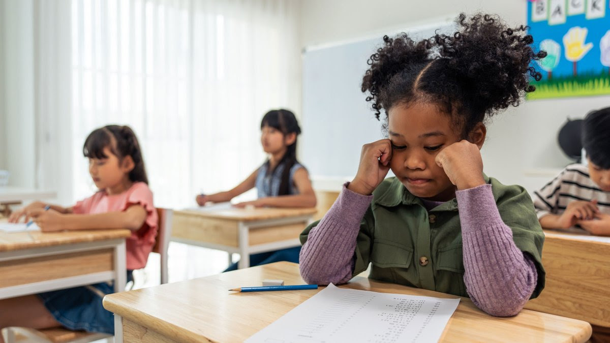 A young black girl sits at her desk in her classroom looking at her paper appearing sad 