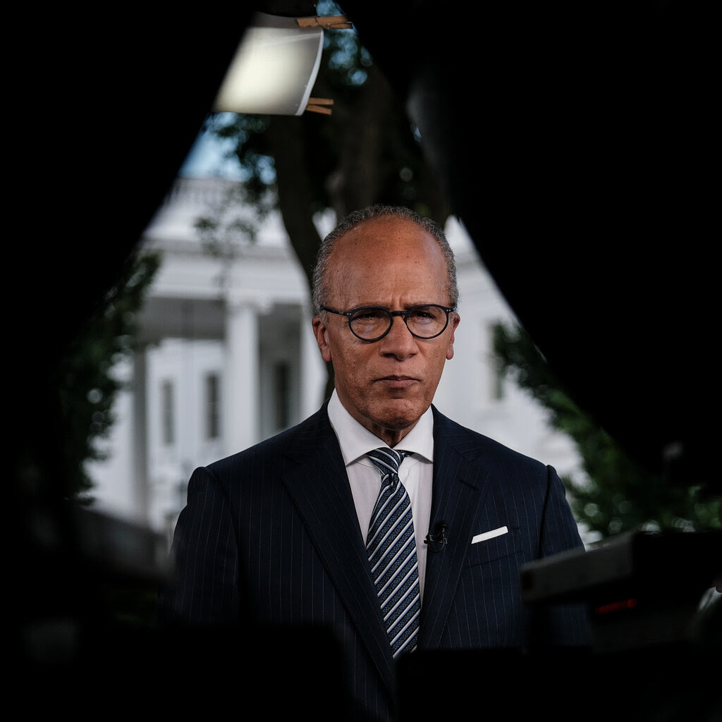 A man wearing glasses, a dark suit and striped tie faces the camera, the White House behind him. 