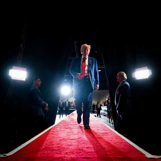 A low-angle shot of Donald J. Trump, wearing a blue suit and a red tie, walking on a red carpet between curtains.