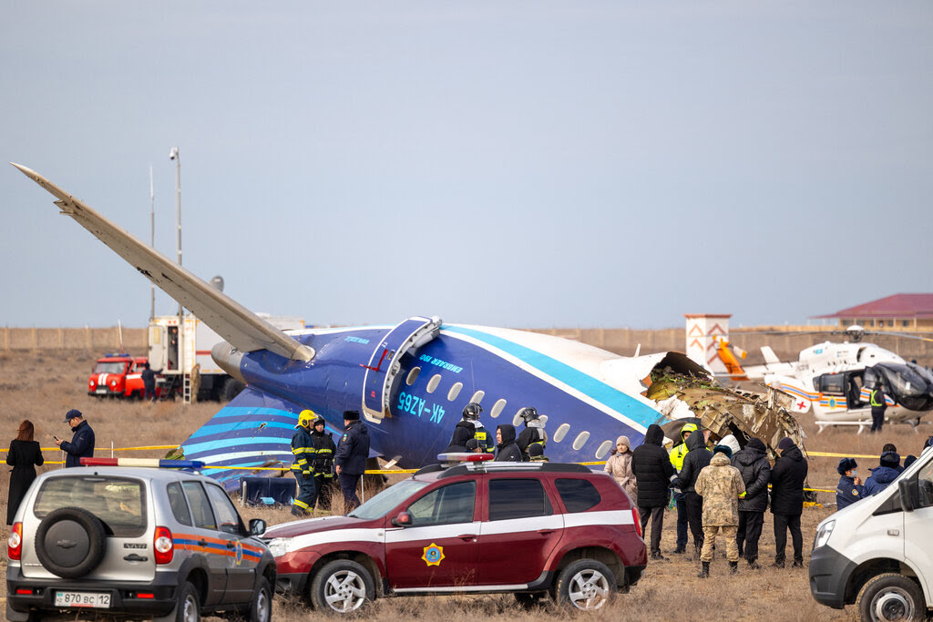 Emergency workers and vehicles are at the site of a plane crash. Part of the plane, which is painted blue and white, is upside down behind them.