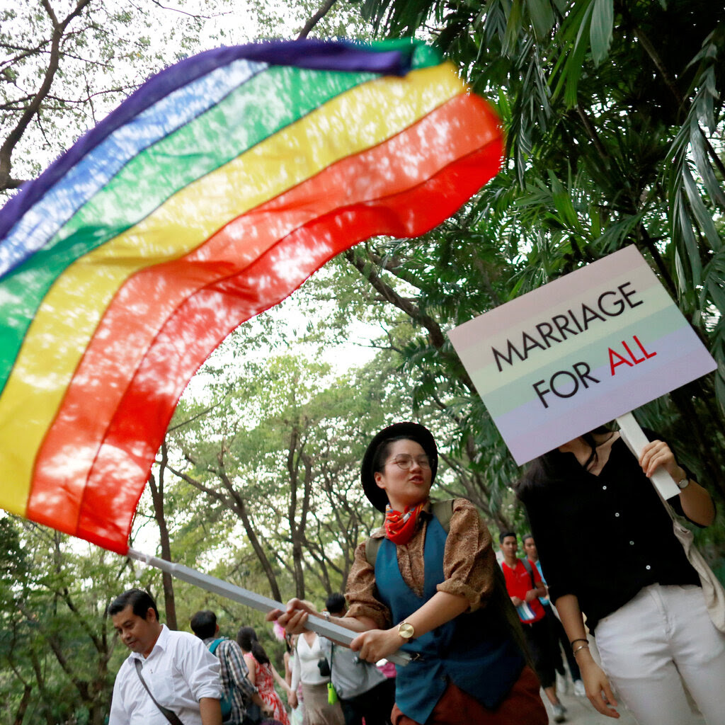 Two people walk along a path in the woods, one waving a rainbow flag and the other holding a sign that reads “Marriage for All.”