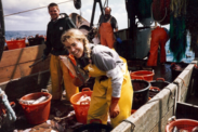 Tonya Wick poses, smiling, aboard a fishing vessel at sea in 1998 in yellow fishing coveralls