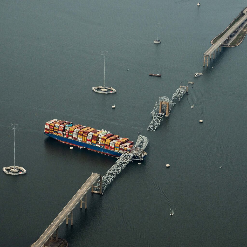 An aerial shot showing a huge cargo ship with its bow trapped under a collapsed bridge span, after the ship slammed into the bridge and it plummeted into the water.