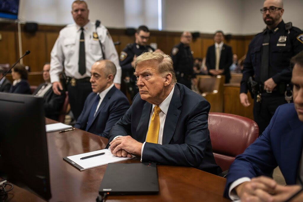 Donald Trump sits at the defendant’s table wearing a dark suit and yellow tie. 