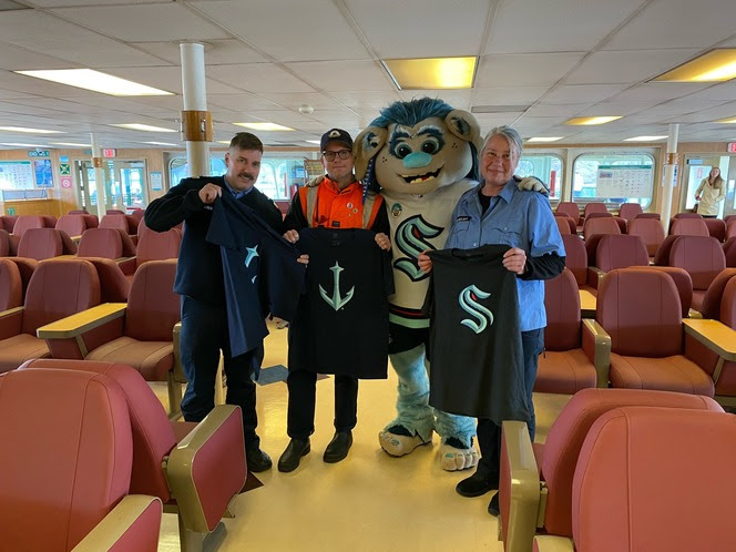 Seattle Kraken mascot Buoy in the passenger cabin of a ferry posing with crewmembers that are holding up Kraken shirts