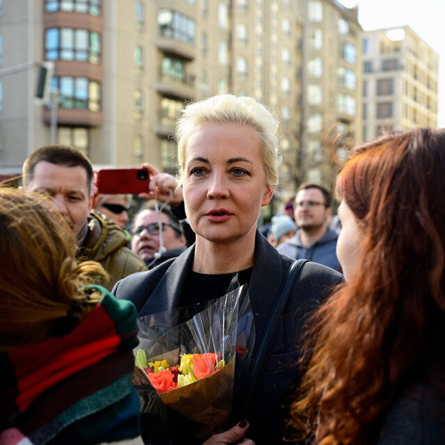 Yulia Navalnaya stands in the middle of a crowd on a street in daylight, a small bouquet of flowers wrapped in paper and plastic and held in front of her.