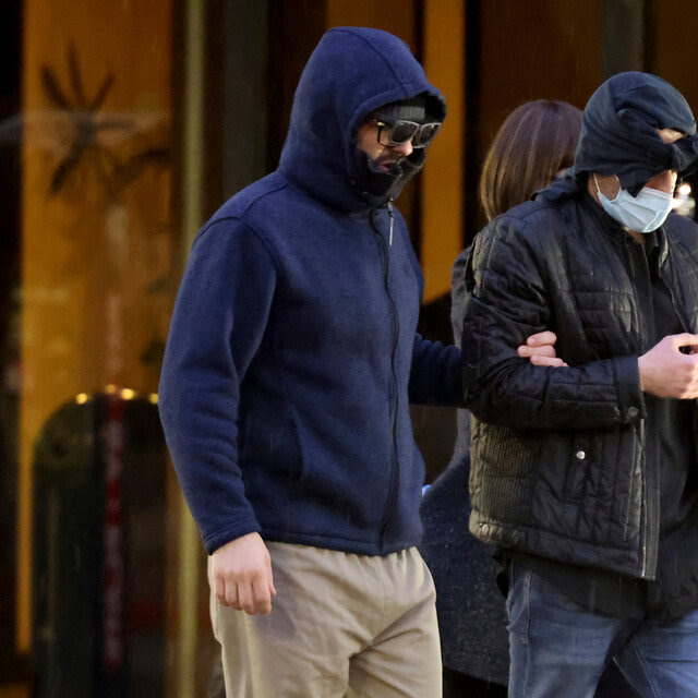 Alexander Smirnov, wearing a blue jacket with the hood up and glasses on, holds onto the arm of another man, dressed in black and with a mask over his face and what seems to be a shirt wrapped around his head.