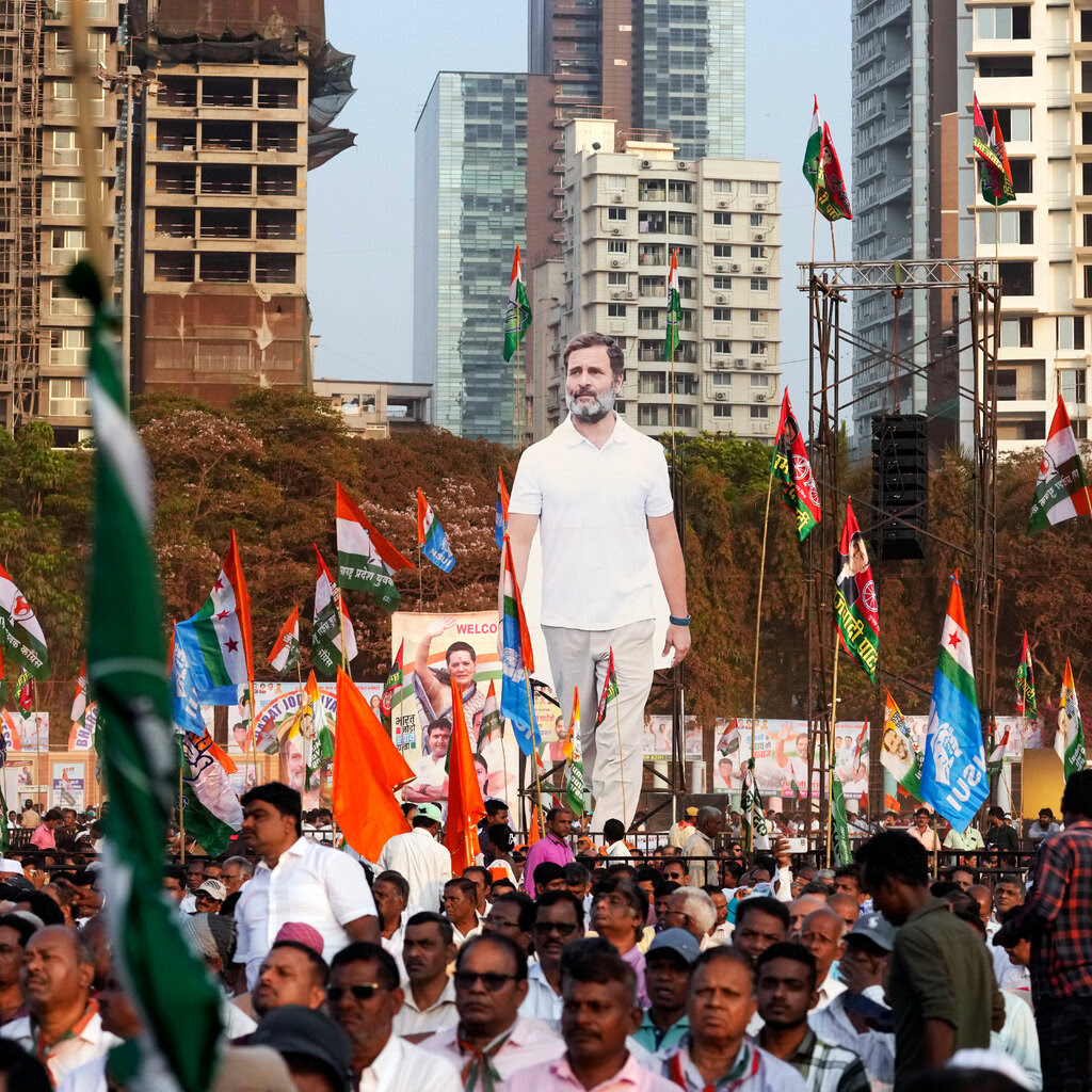 Large cutouts of Rahul Gandhi and other opposition figures towering over a dense crowd near high-rise buildings.