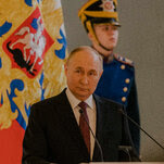 Vladimir Putin, wearing a dark suit jacket and maroon tie, stands in front of two microphones, with a uniformed guard and a flag behind him.