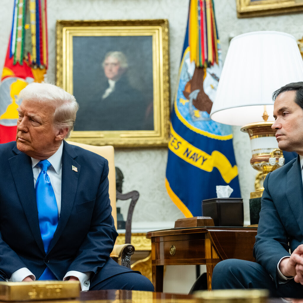 Two men in suits sit in a room with ornate decorations.