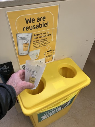 A yellow recycling bin with a poster about returning reusable cups and a hand holding a reusable cup above it