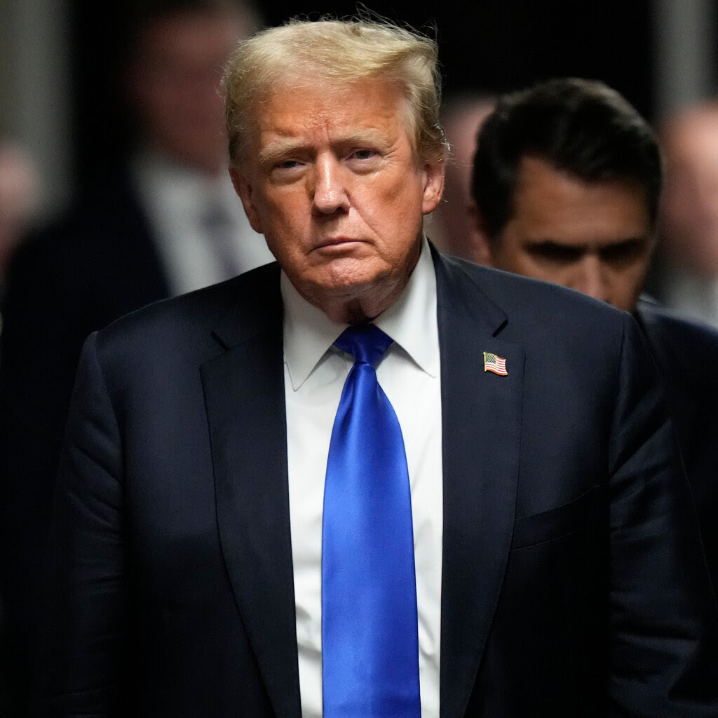 Donald Trump looks somber in a suit with a blue tie and an American flag. 