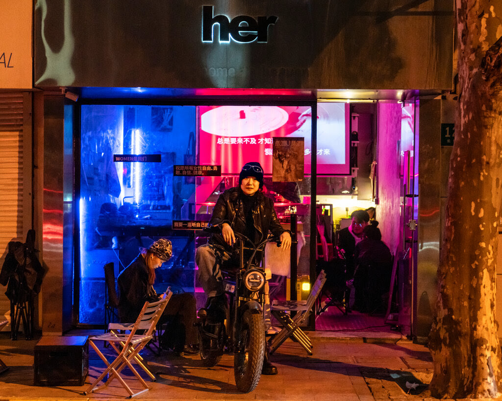 Du Wen, wearing a black beanie, a black leather jacket and black pants, sits on a motorbike outside a bar lit in neon colors at night. Another woman sits on a chair next to her on the sidewalk.
