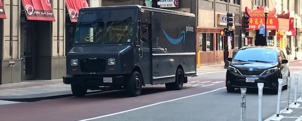 a picture of a boston street with a bus lane,
a travel lane, and a separated bike lane. there is an amazon delivery
truck parked illegally in the bus lane