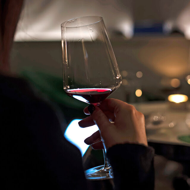 A view over the shoulder of a person holding a glass of red wine in a moodily lit restaurant.