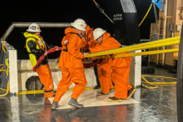 Four crew members in orange jumpsuits work on the deck to retrieve a trawl at night during rough weather