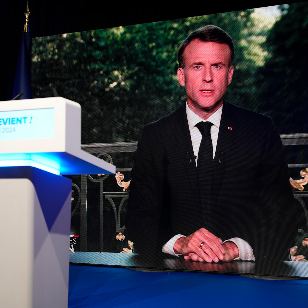 A television screen shows President Emmanuel Macron of France in a dark suit and tie. 