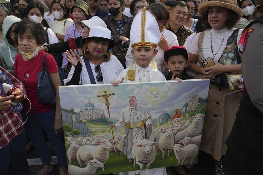 A crowd of people gathered for Pope Francis's visit. At the front of the group is a child holding a painting of Saint Francis with a flock of sheep.