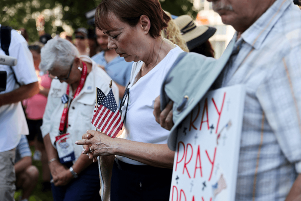 Middle-aged white woman in prays with eyes closed and hands holding two American flags. She is in a crowd of people praying. 