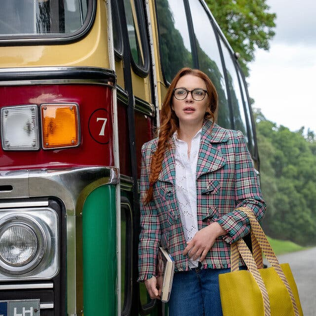 A woman in a plaid jacket holding a yellow tote bag stands next to a colorful bus on a country road.