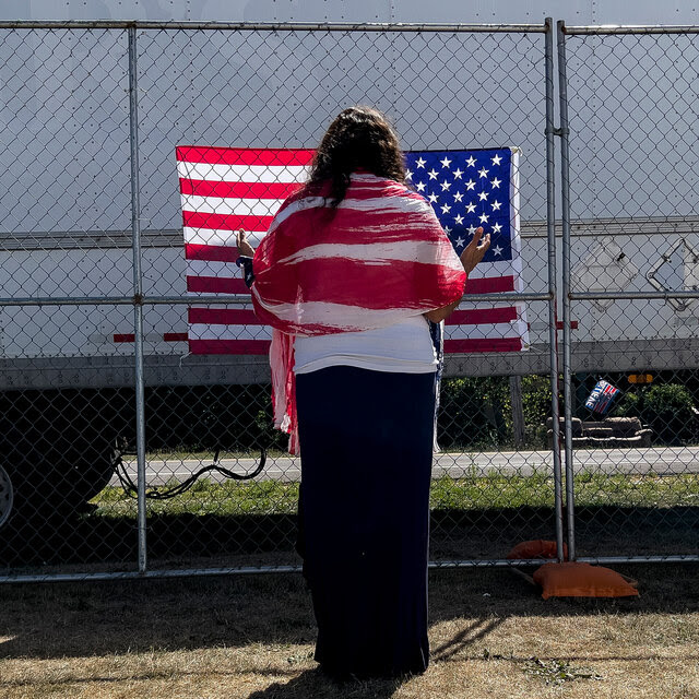 A woman wearing a white and red shawl prays as she faces an American flag on a chain-link fence.