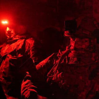 Red flashlights illuminate a dark room in which two soldiers wearing combat gear wait.