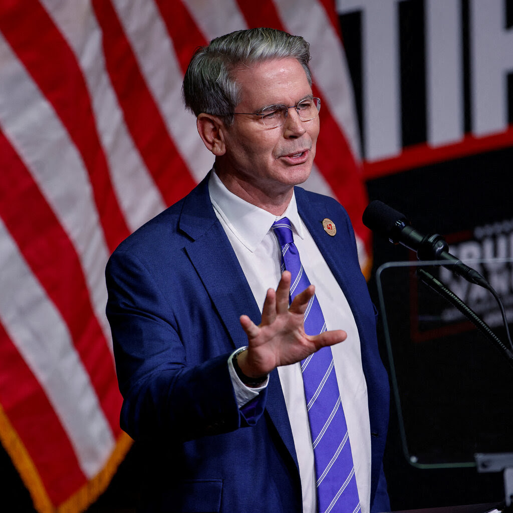 Scott Bessent, wearing a blue suit jacket, white shirt and blue and white striped tie, gestures as he stands behind a lectern with a Trump-Vance sign on it.