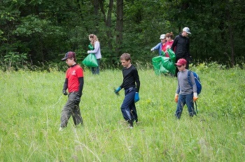 three young boys in jeans and long-sleeve shirts walk through grassy area with bags, while adults behind them pull invasive plants