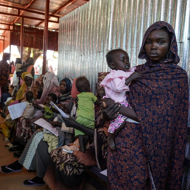 A Sudanese woman holds an infant near a line of other women with children against a wall.