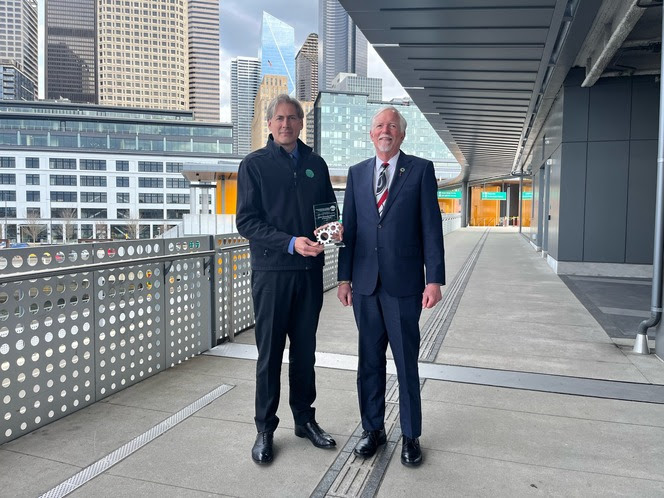 Two people posing for a photo at Colman Dock in Seattle with one holding an award